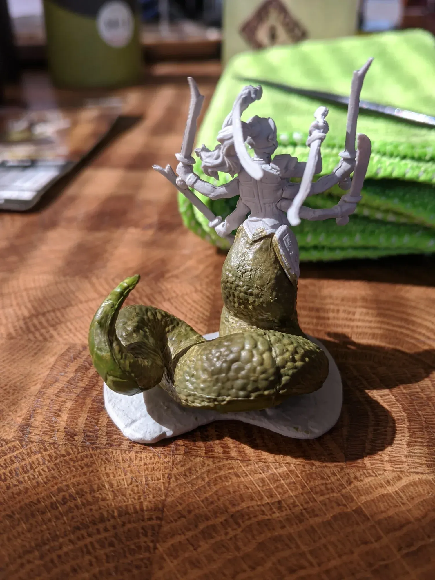 A partially painted tabletop game miniature of a snake-bodied woman with six arms holding swords, on a wooden table.