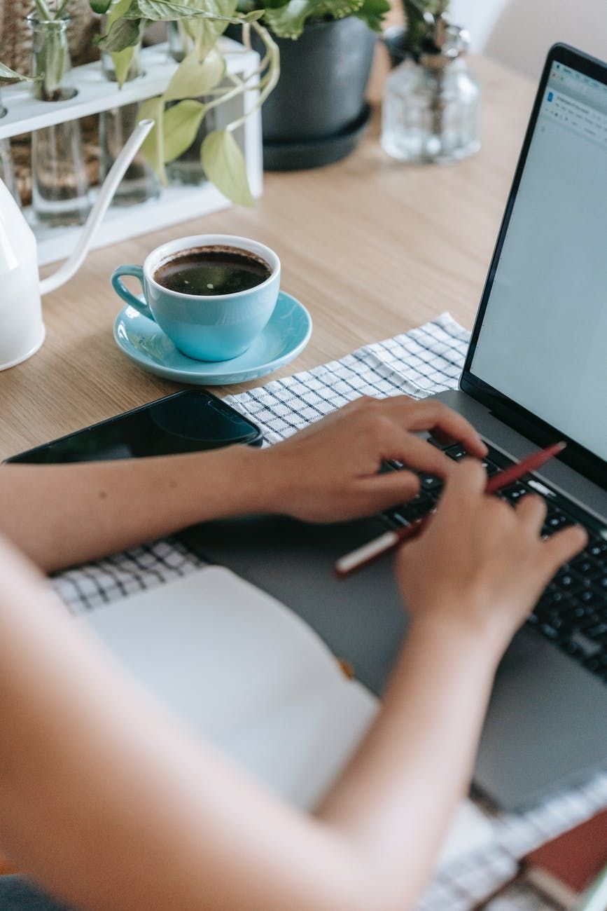 Person typing on laptop, coffee cup, and notebook on a wooden table with plants in the background.