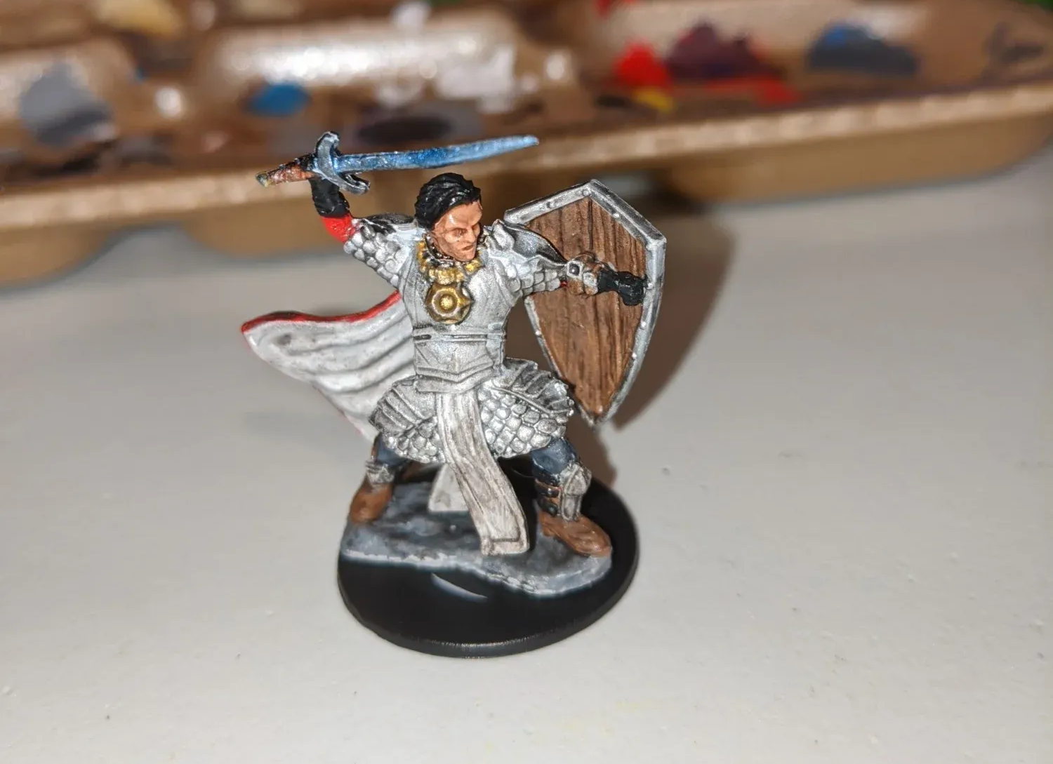 Miniature figure of a knight, painted with a blue sword, white cape, brown shield, and holding a sword aloft.