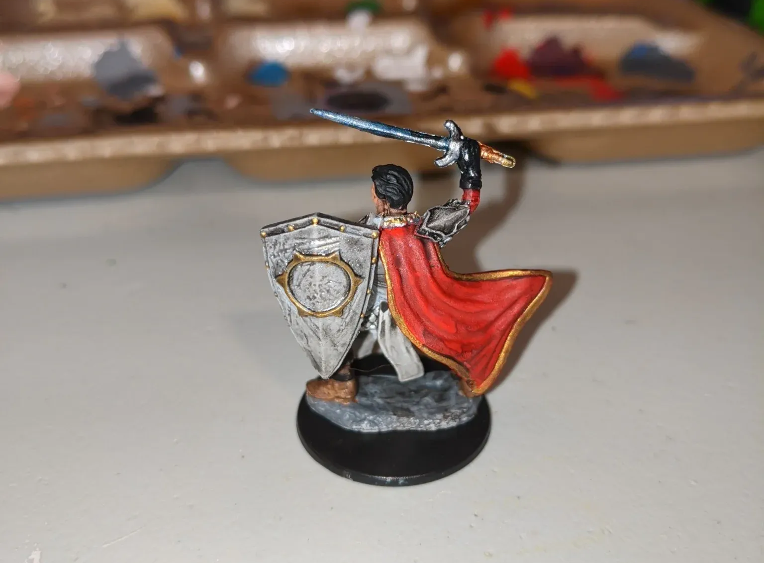 Miniature figurine of a knight in red cape holding a sword and shield on a black base.