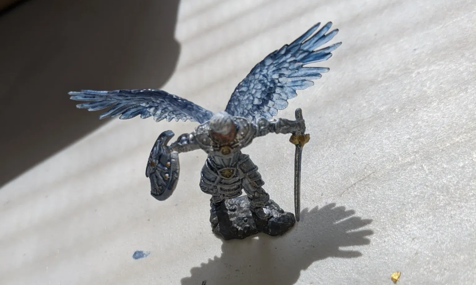 Miniature angel with blue-grey wings, holding a sword and shield, on a white surface.