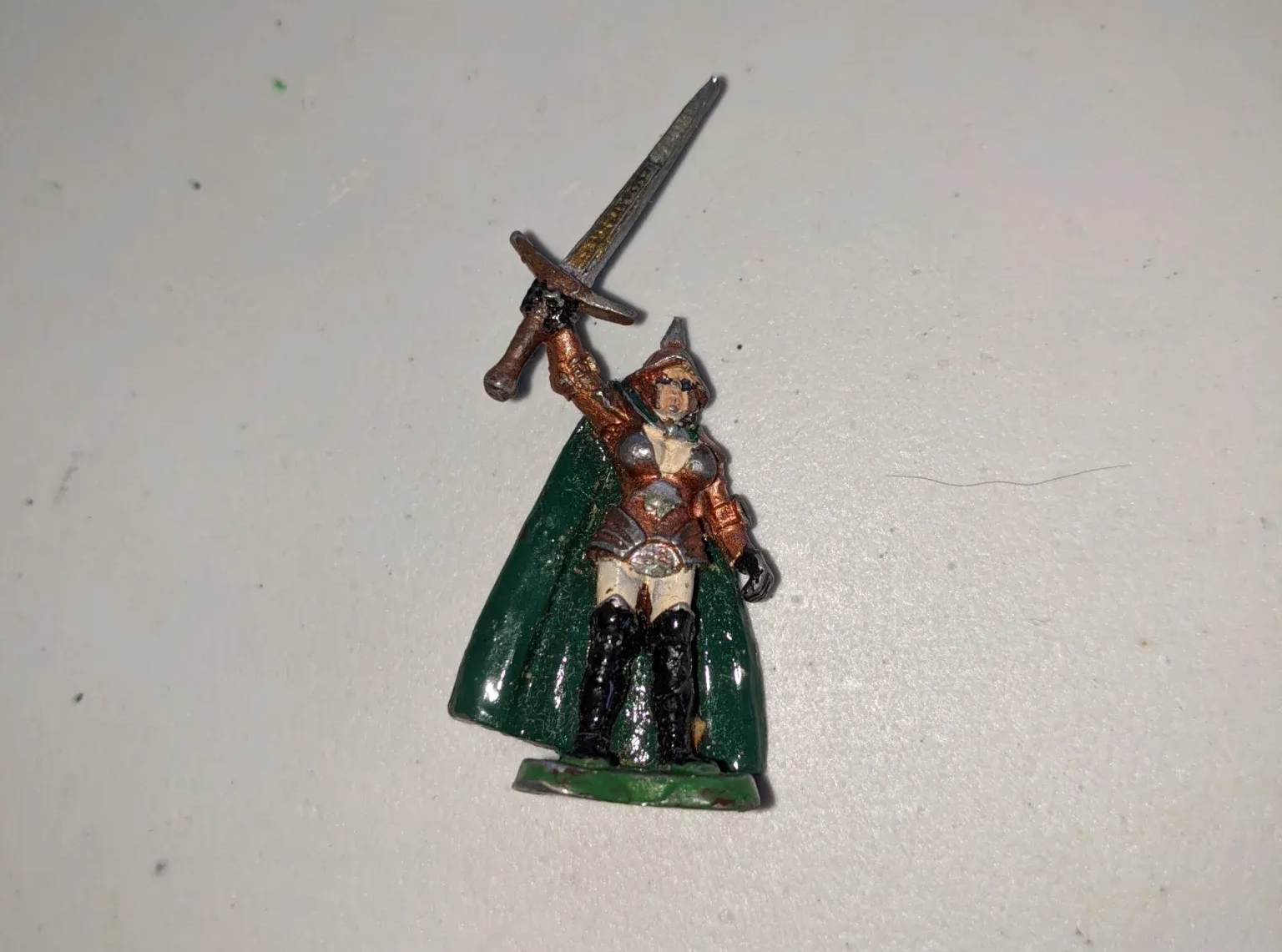 Miniature figure of a warrior in green cape raising a sword, on a white surface.