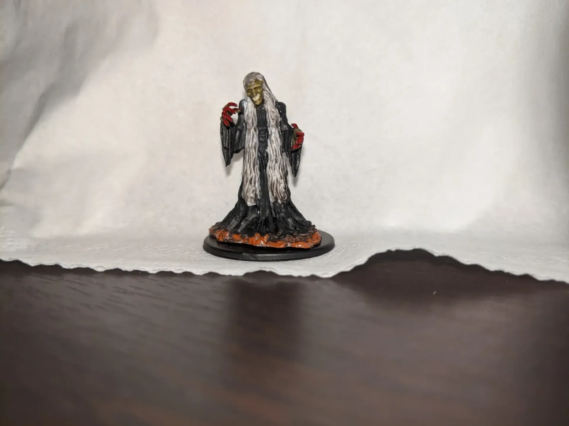 Miniature of a robed, skeletal figure with long white hair, holding a red item; on a wood table.