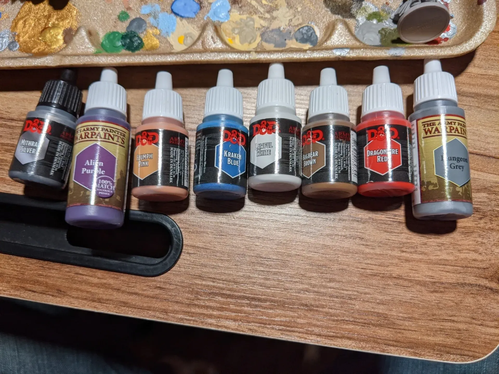 Paint bottles of various colors, arranged on a wooden surface. A palette with paint is in the background.