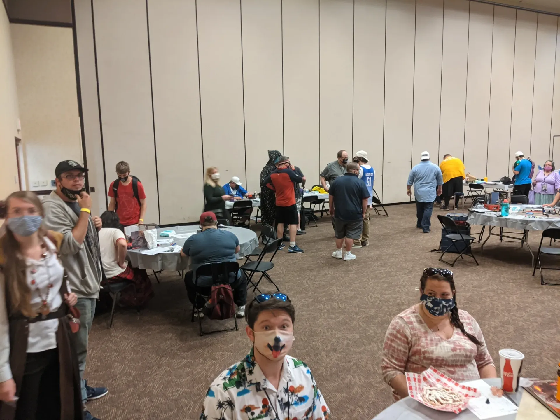People gather at a convention, some wearing masks, tables set up, neutral-colored walls.