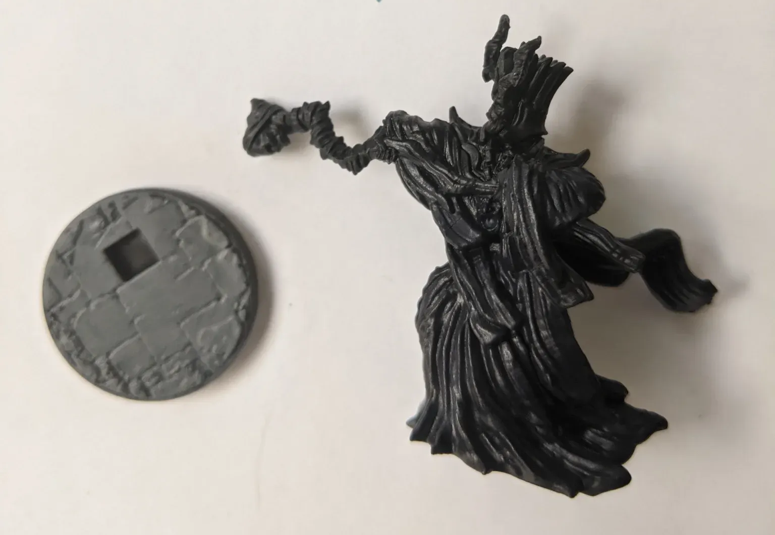 Black fantasy miniature next to a grey round base. The miniature depicts a cloaked, shadowy figure reaching out.