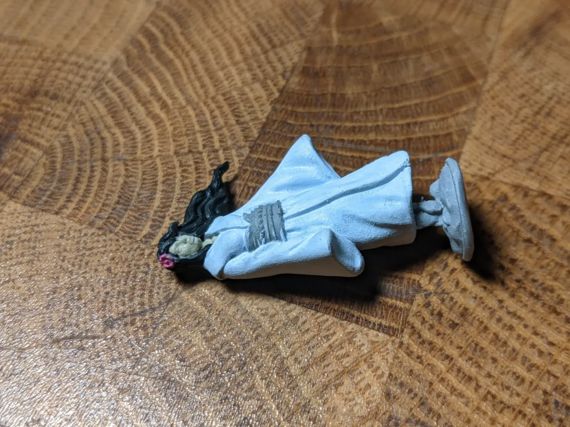 Miniature figure of a person in white robe, black hair, lying on a wooden surface.