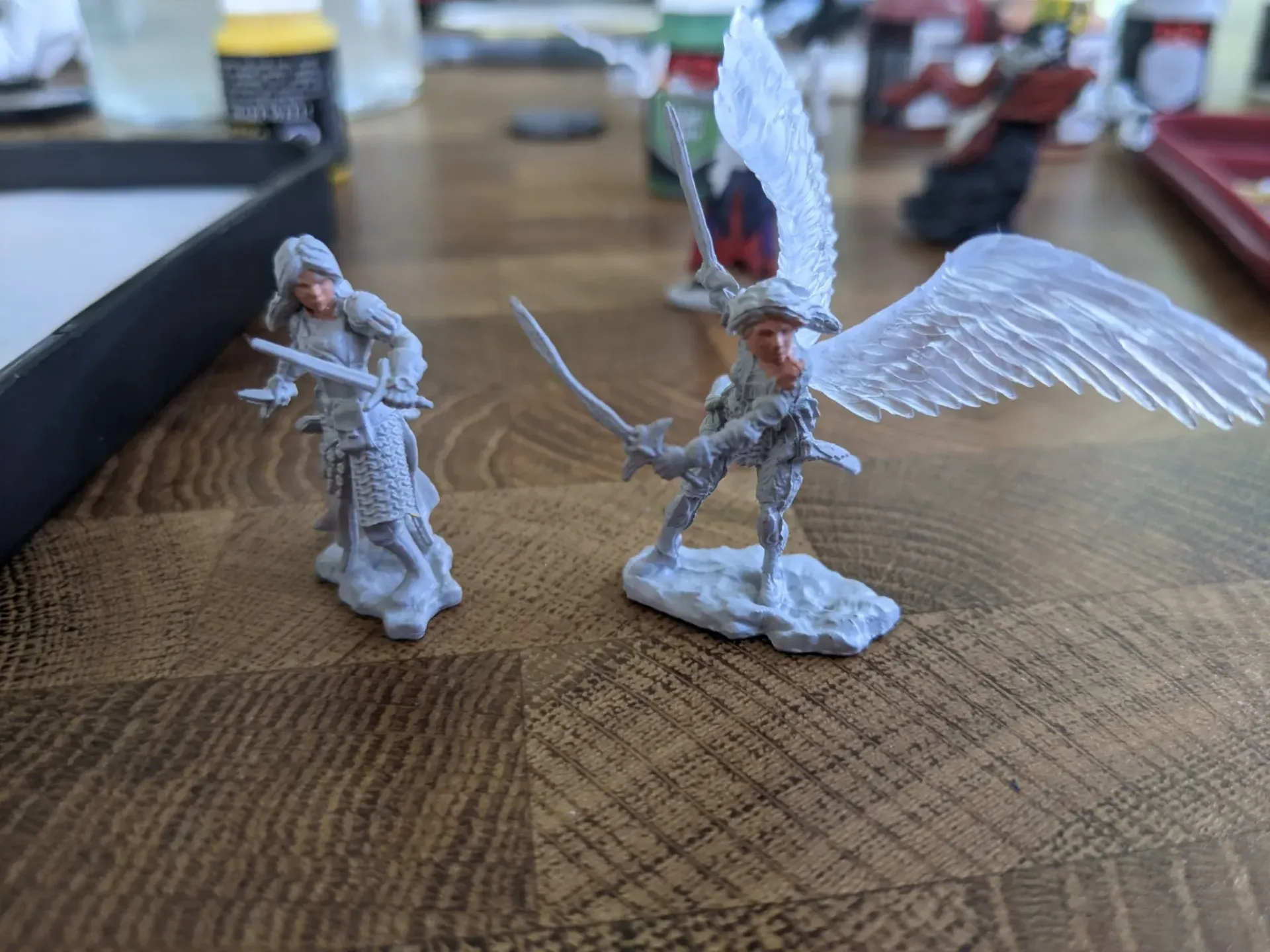 Two unpainted miniature fantasy figures on a wooden surface. One is a winged woman, the other is a standing woman with a sword.