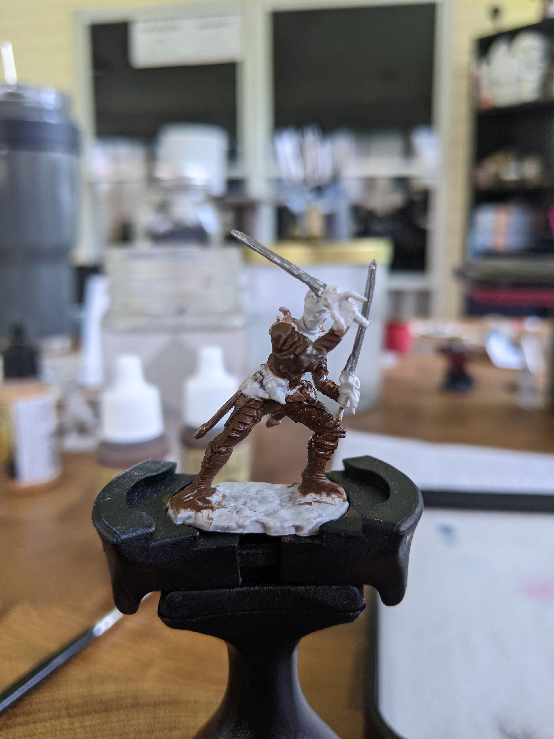 Miniature figure, brown armor and silver swords, on a painting stand. Light tan base, hobby area background.