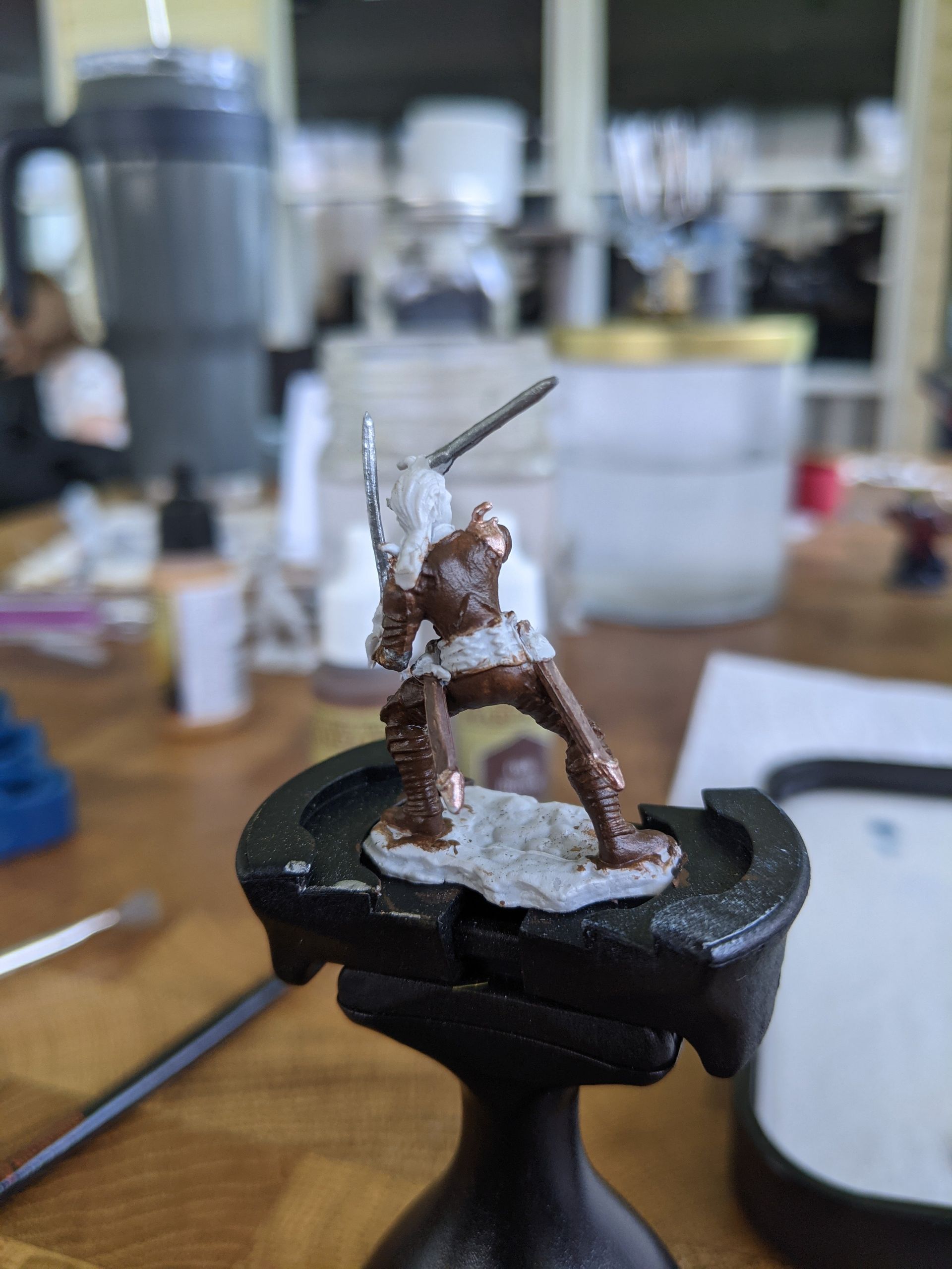 Miniature figure being painted, brown armor, white base, on painting handle, interior.