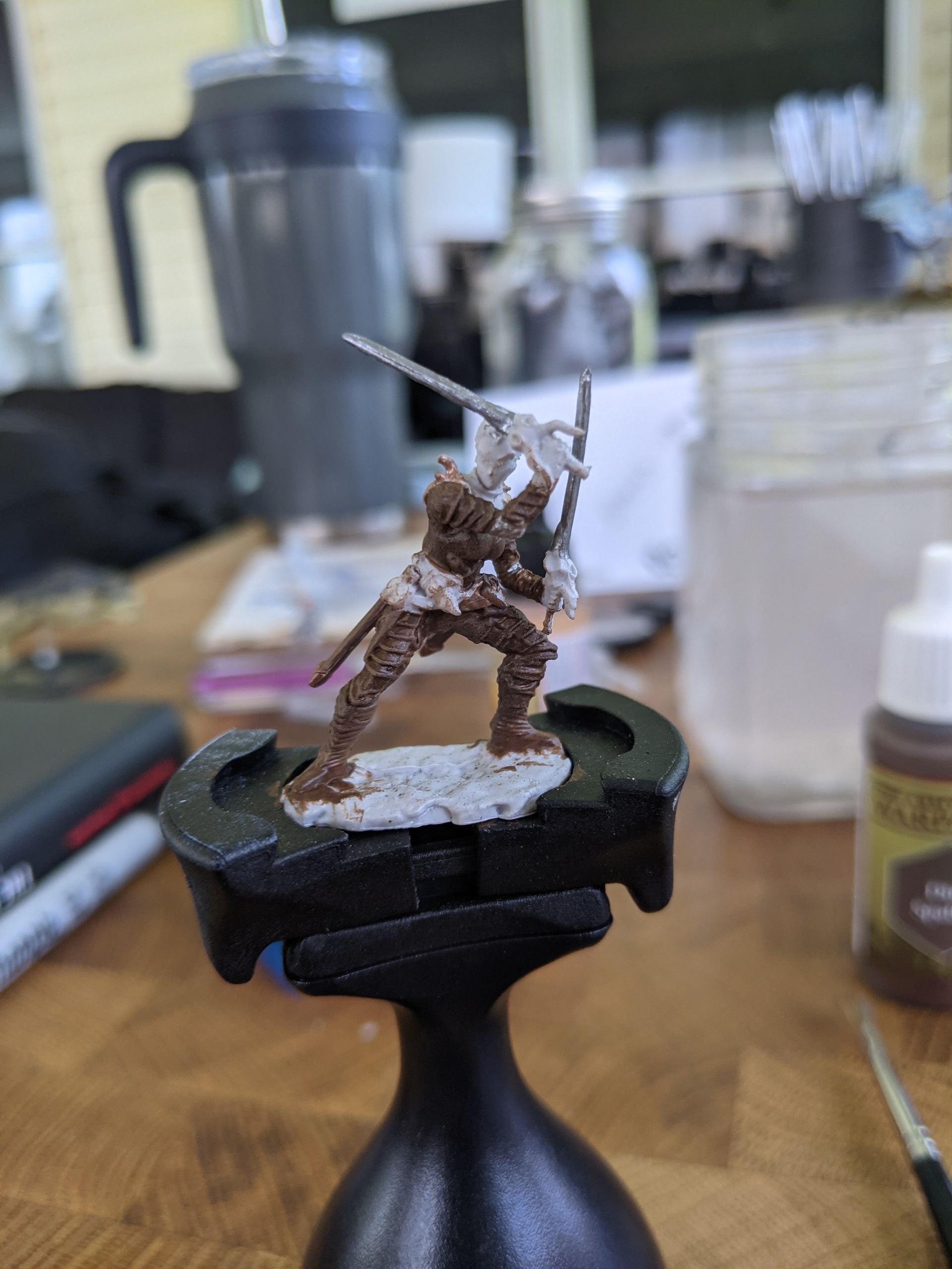 Miniature figure of a warrior in brown armor, held in a painting stand. Wooden table in the background.