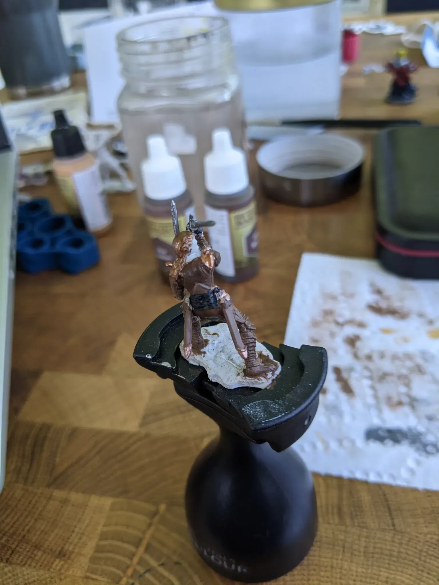 A miniature figure, painted in shades of brown, perched on a painting handle. The setting is a messy table.