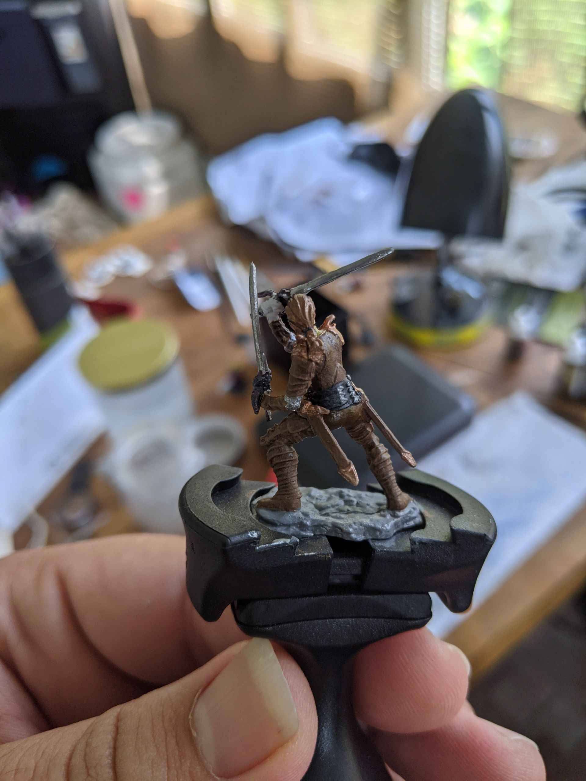 Miniature figurine of a warrior with two swords. Brown armor, standing on a textured base, held in hand.