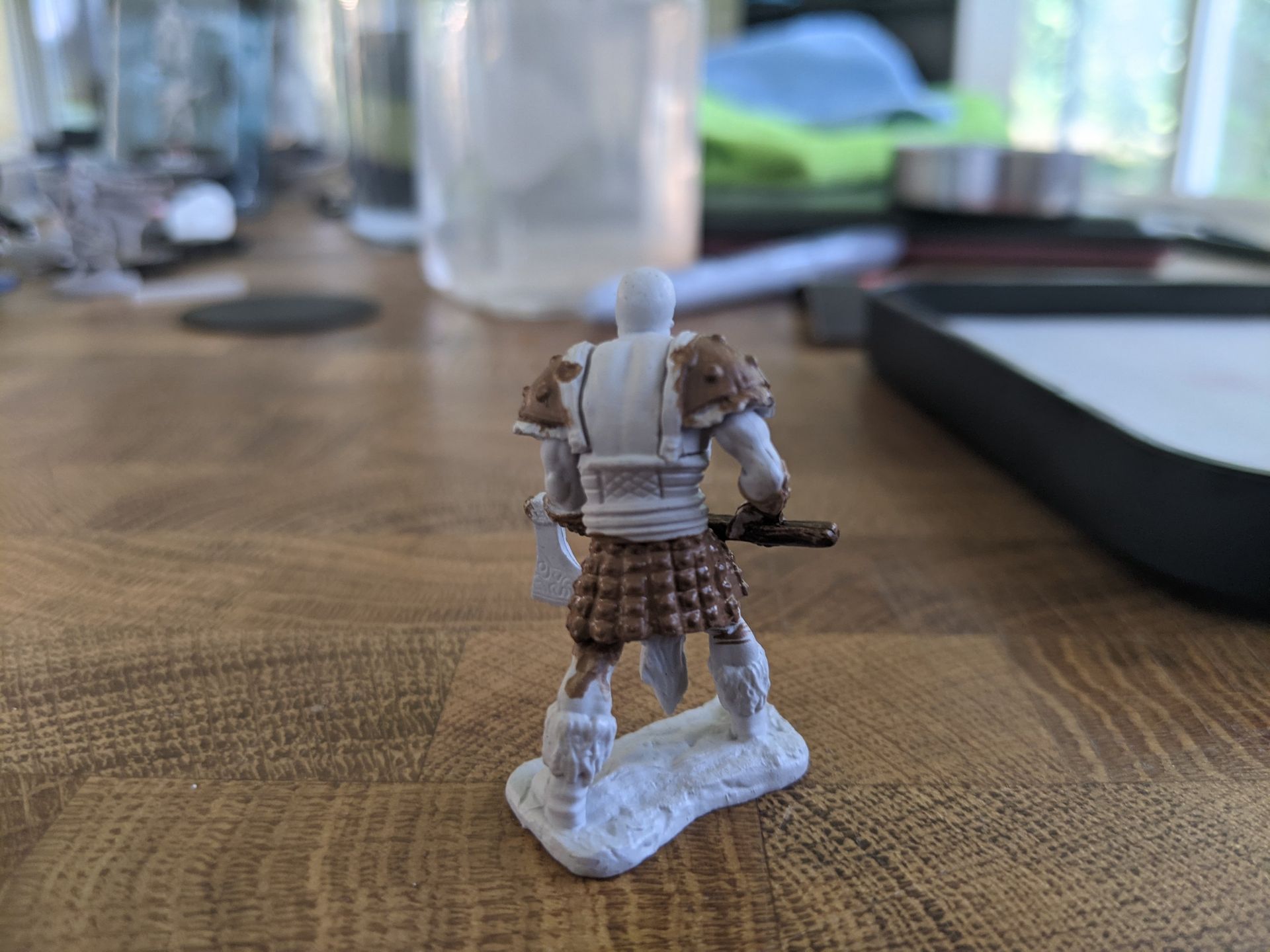 Miniature figure of a warrior, back view. Light grey armor, brown kilt, holding a weapon. White base on wooden surface.