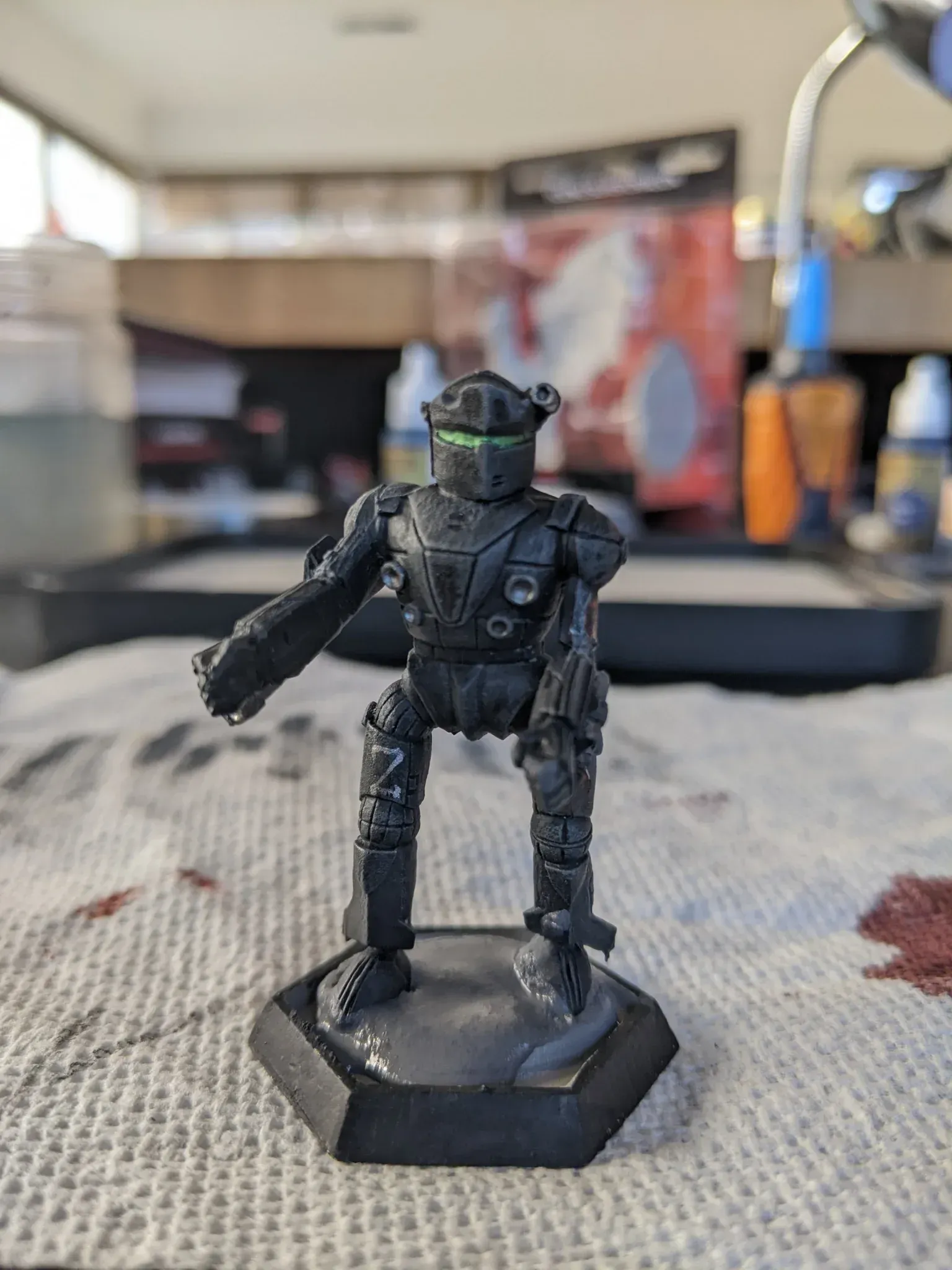 A miniature, black robot figurine on a hexagonal base, with a glowing green visor.