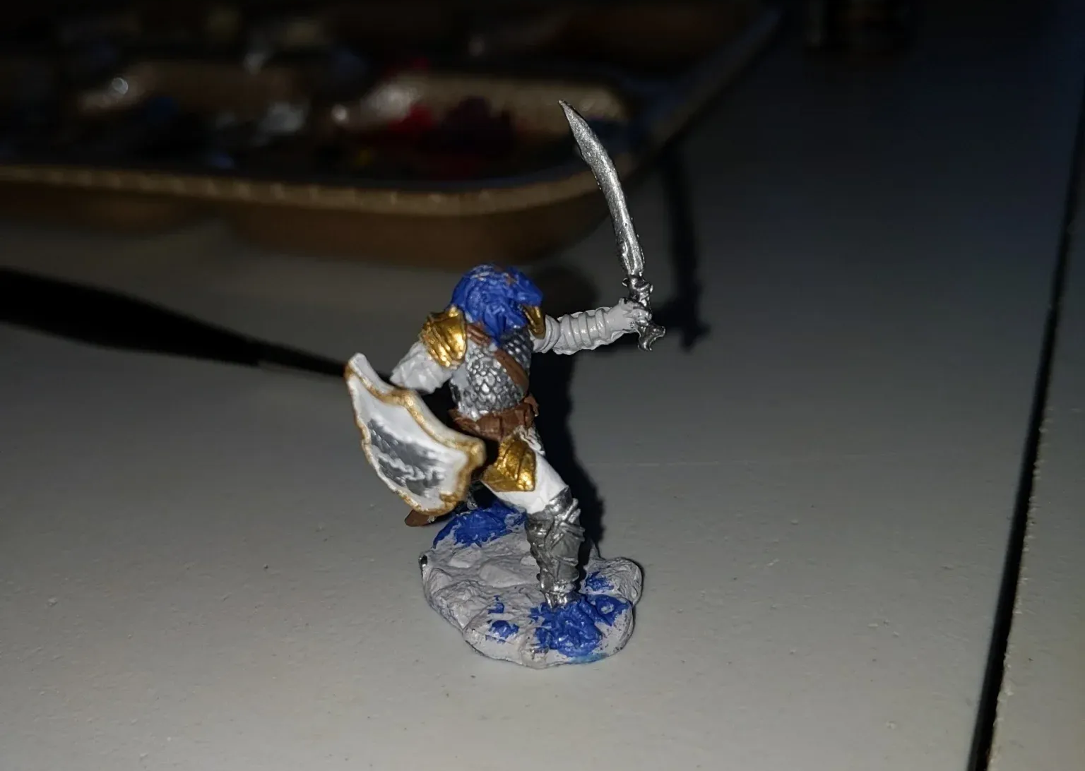 Miniature figure of a warrior in blue and gold armor, sword raised, on a white surface.
