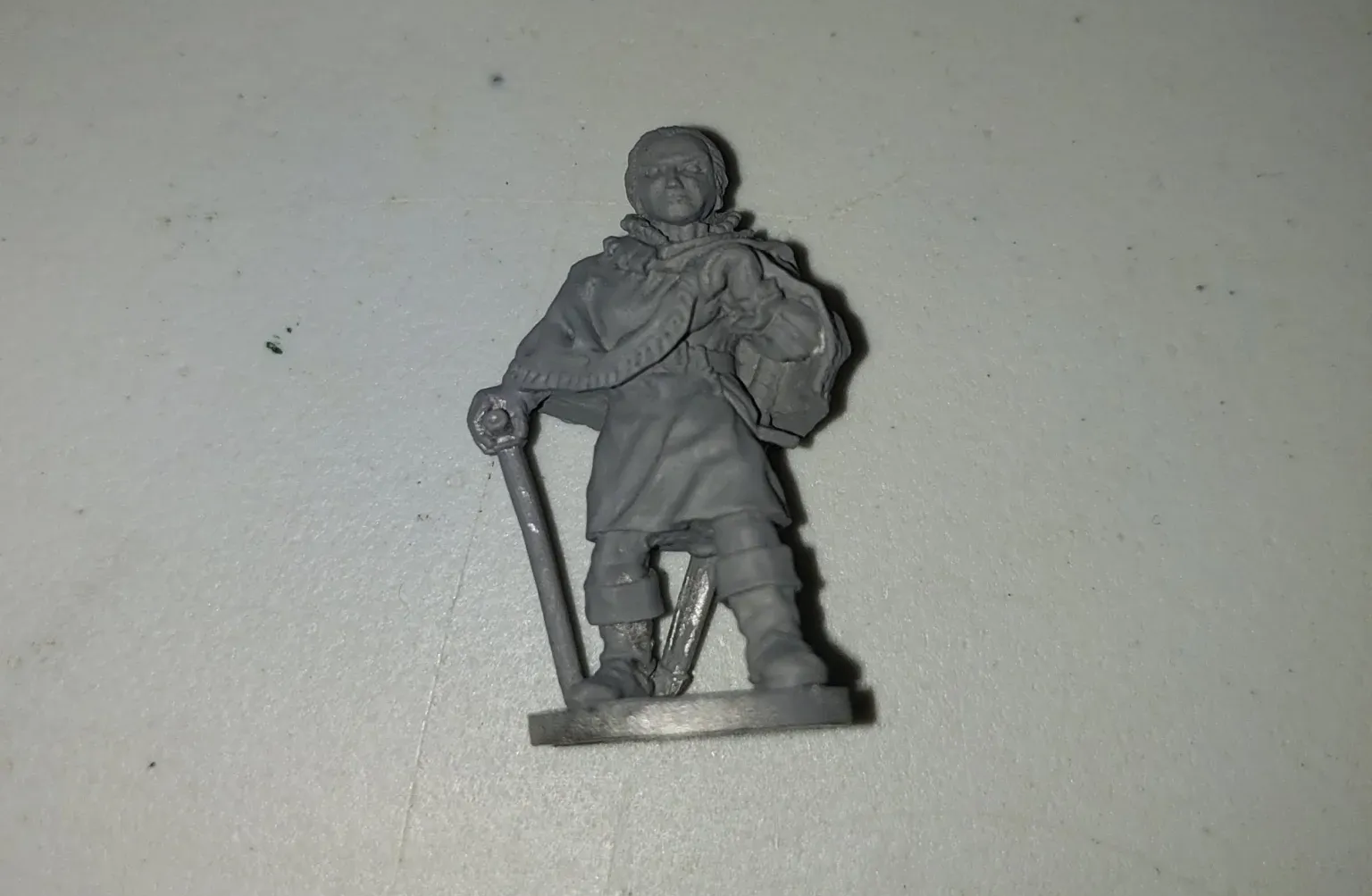 Miniature figurine of a man wearing a cloak, holding a staff and a bag, standing on a base.