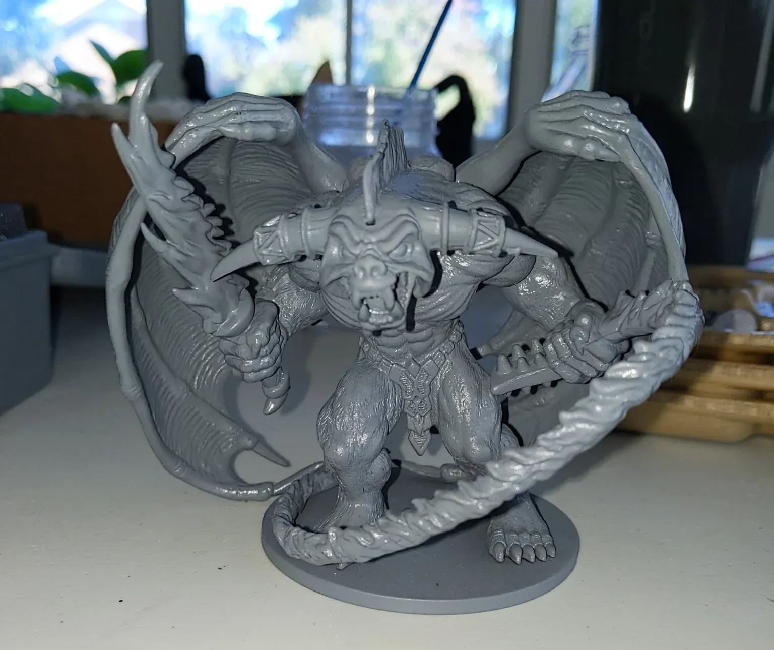 A grey plastic demon figurine with wings and multiple heads, posed on a white surface.