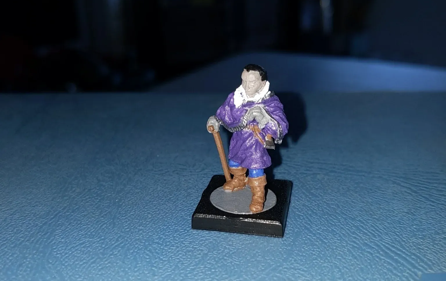 Miniature figure of a man in purple robes, holding a cane, standing on a small black base.