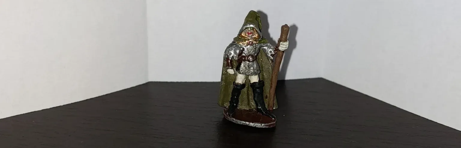 Figurine of a wizard with a staff, green cloak and hat. It is on a dark surface with white walls.