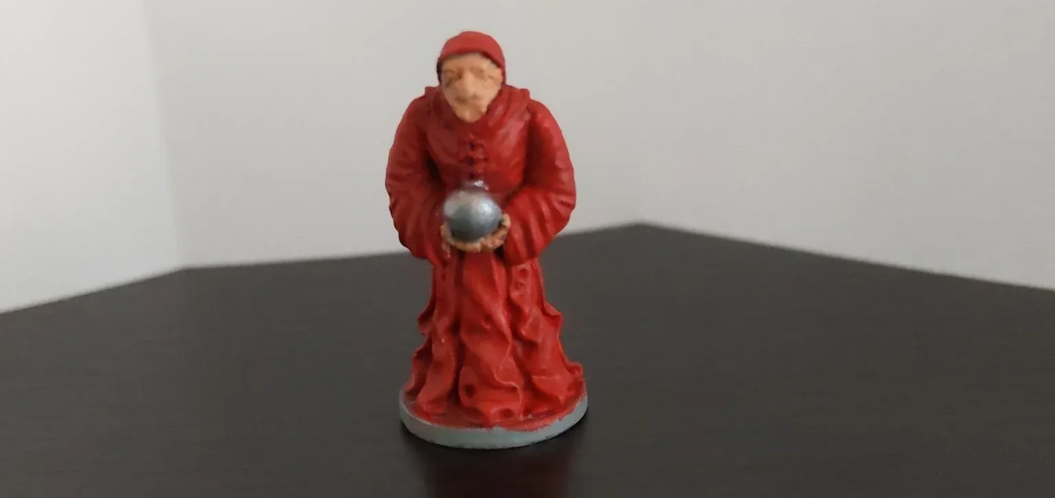 A red robed figurine, holding a silver ball. It's on a dark surface, near a white wall.