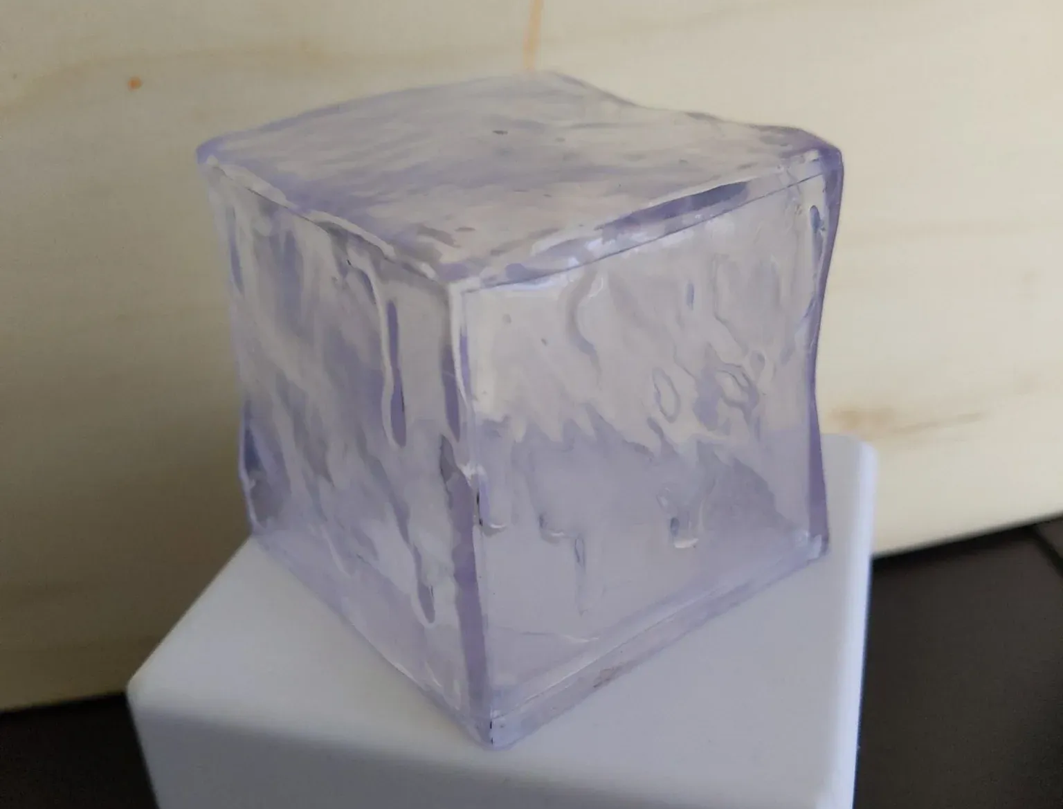 Clear ice cube on a white block against a light-colored wooden background.