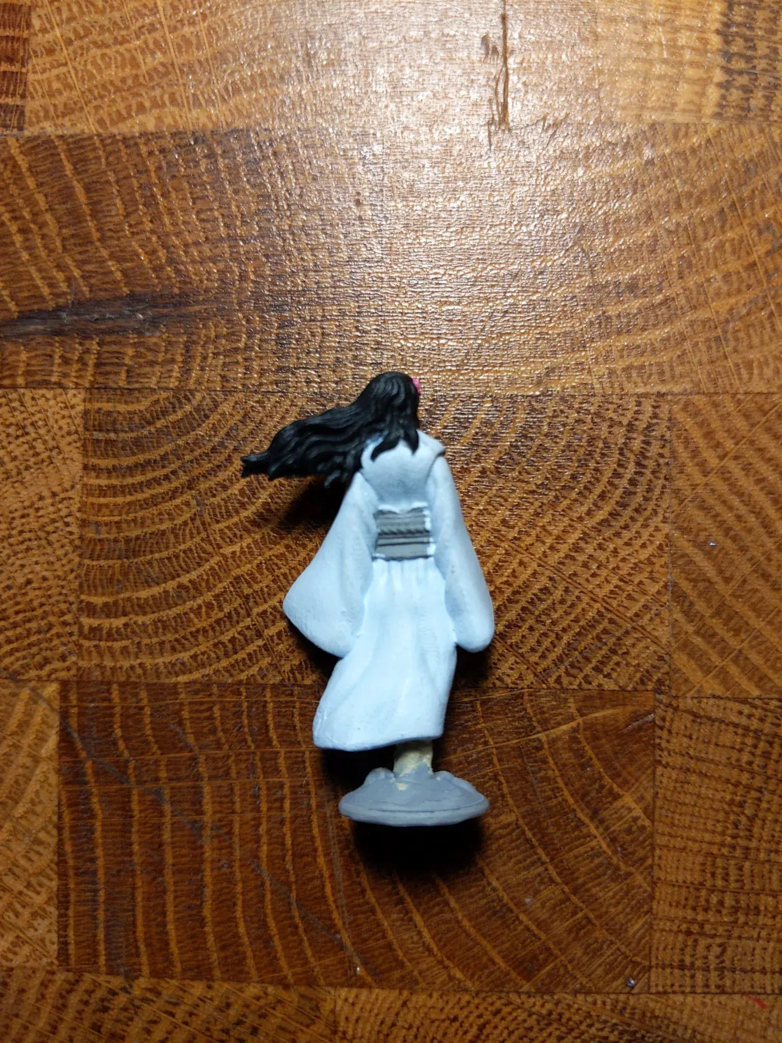Miniature figure of a woman in a white robe, facing away, on a wooden surface.
