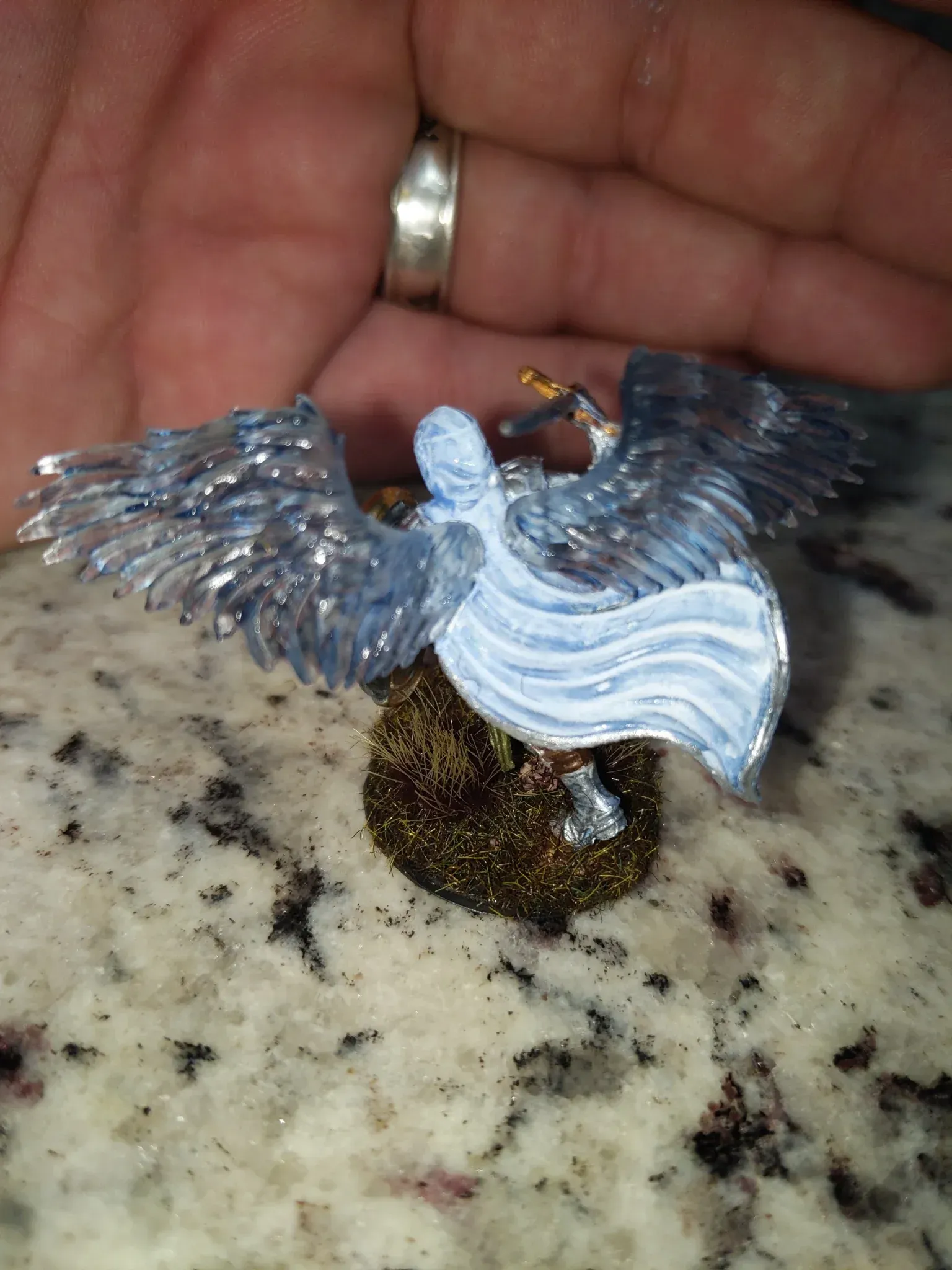 A miniature, silver angel with outstretched wings, on a bed of brown sparkle, held in a person’s hand.