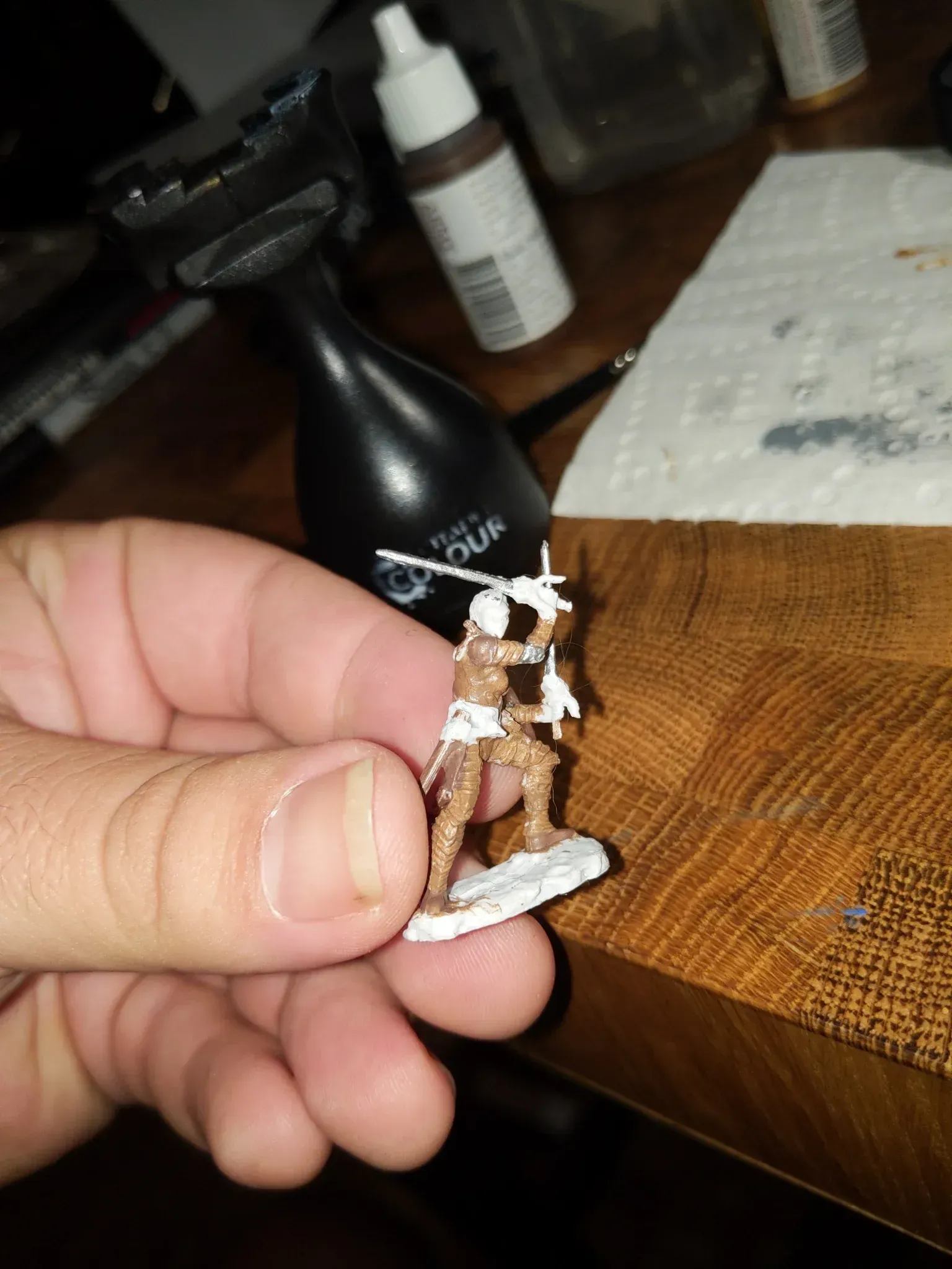 Hand holding a miniature figurine of a warrior with swords, painted brown and white, on a white base.