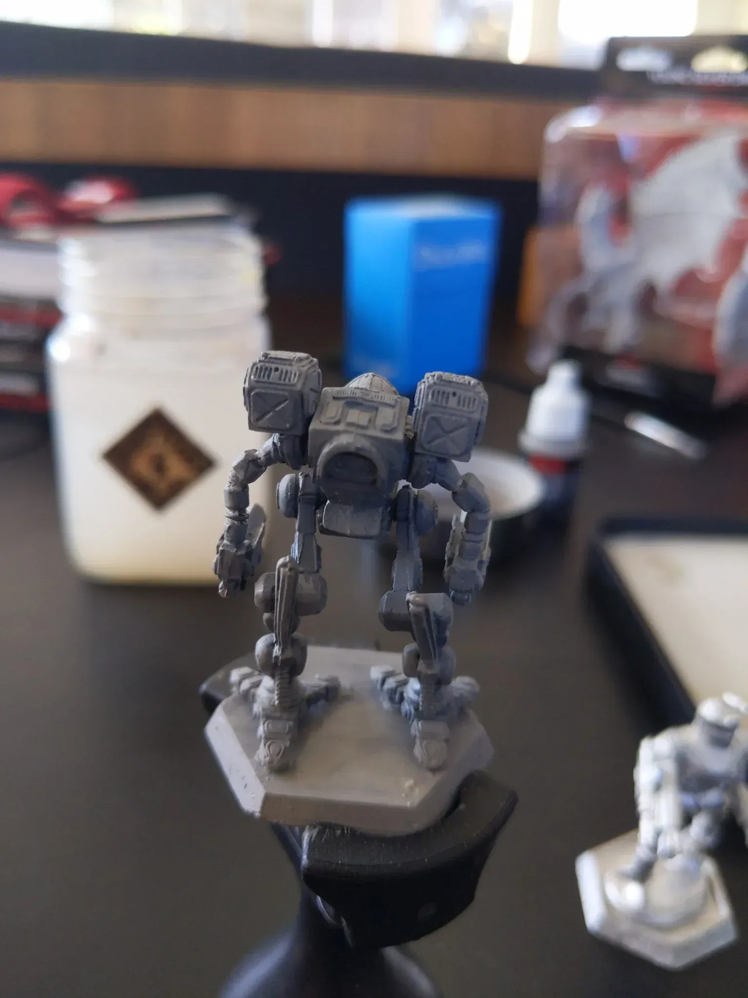 Miniature tabletop game mech figurine, primed gray, on a black base, with painting supplies in background.