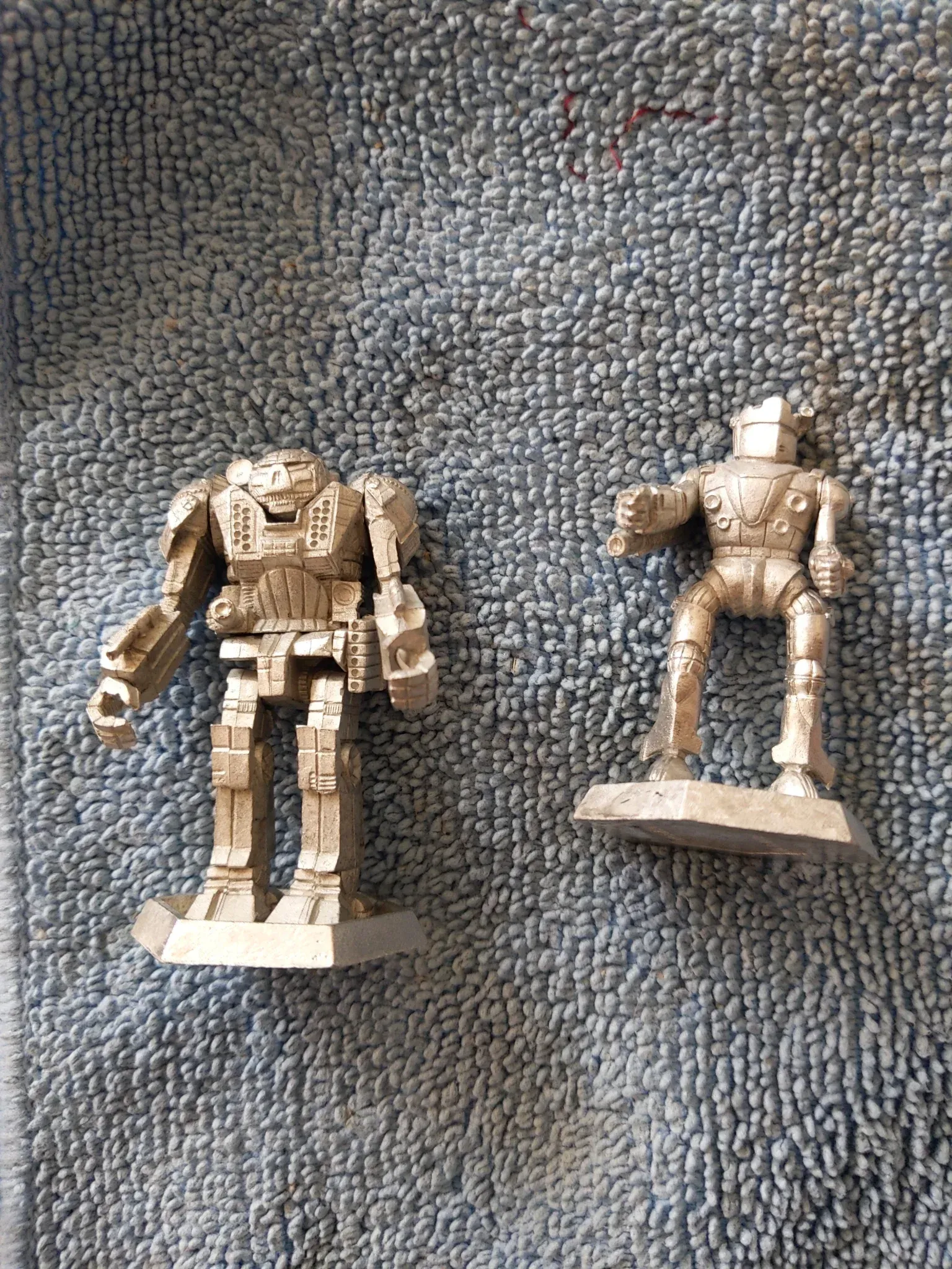 Two silver robot miniatures on a textured surface. One is a larger mech, the other is a smaller humanoid robot.