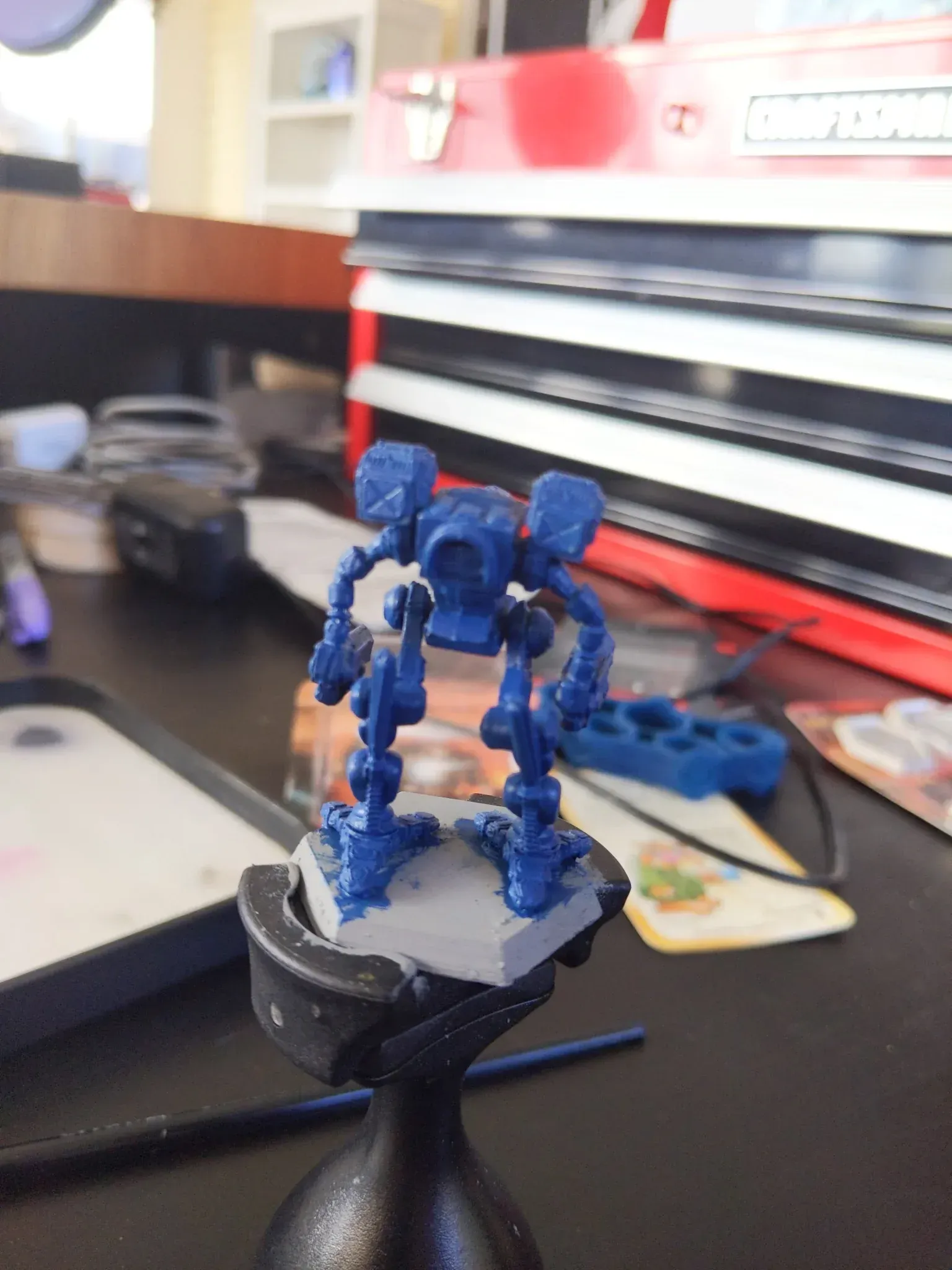 A blue miniature robot figure, painted on a gray base, sits on a black painting stand with tools in background.