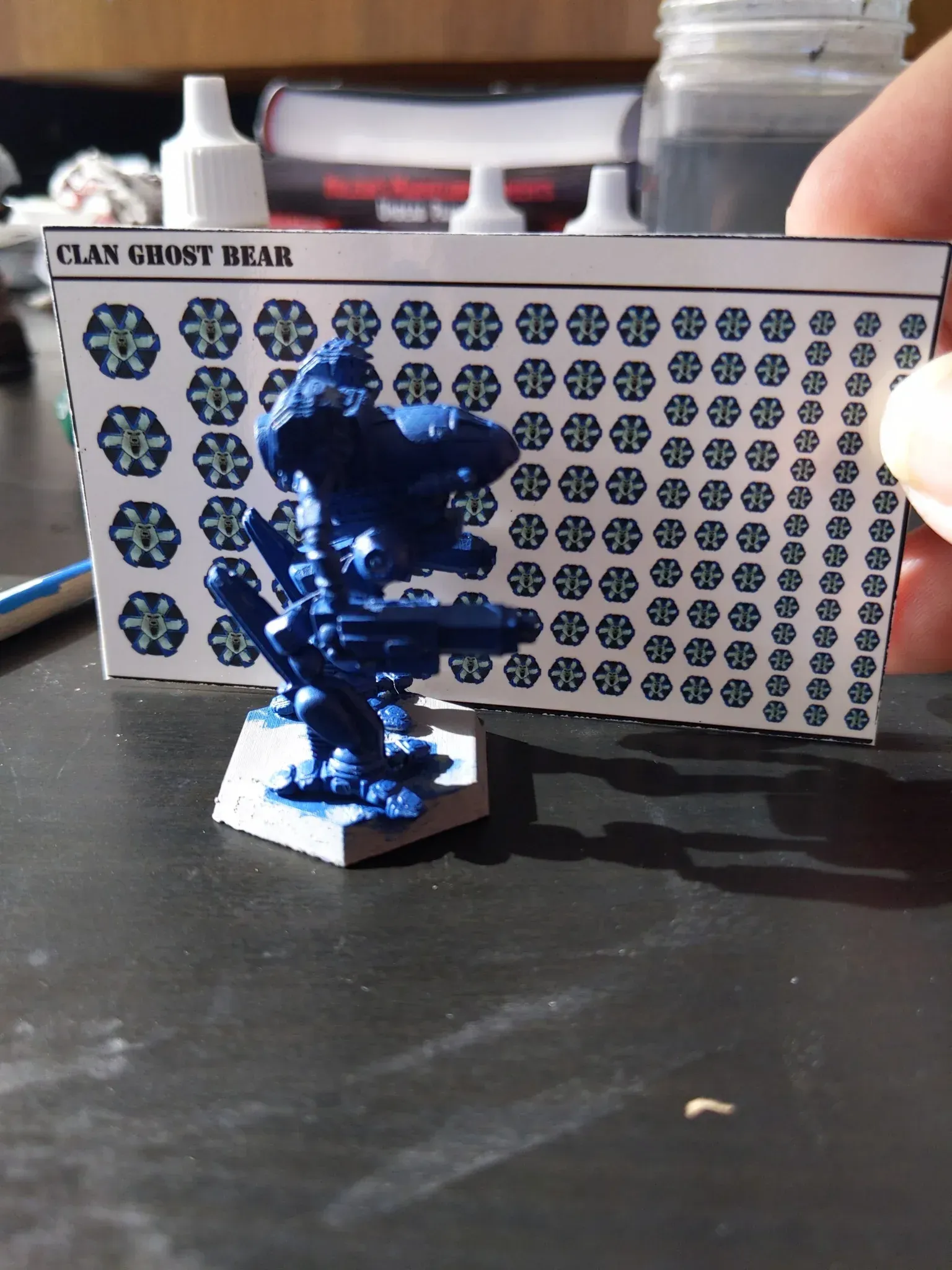 A blue miniature robot with Clan Ghost Bear decals, held near a sheet of matching decals.