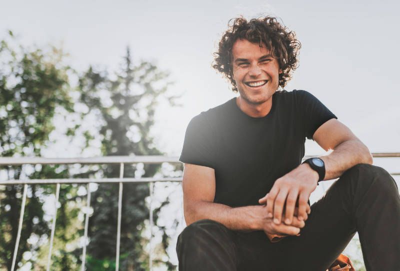 A young man is sitting on a railing with his legs crossed and smiling.