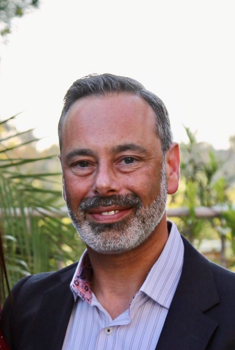 Middle-aged man with salt-and-pepper beard smiles outdoors, wearing a suit jacket over a striped shirt.