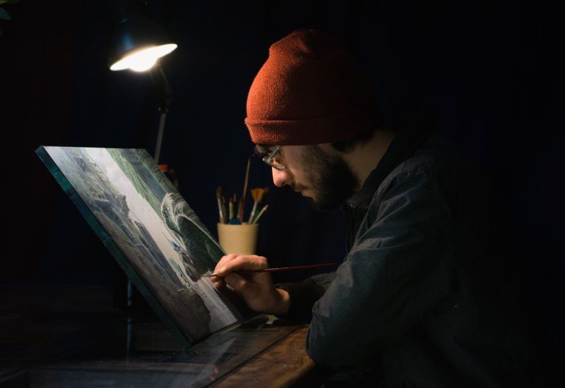 A man in a red hat is painting a picture in the dark.