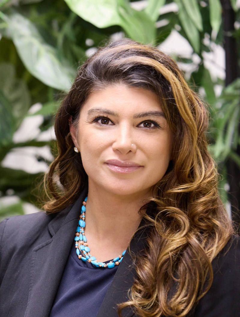 Woman with brown hair, wearing a blue necklace and blazer, smiles in front of foliage.