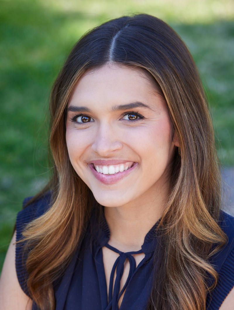 Smiling woman with dark hair and highlights against a blurred outdoor backdrop.