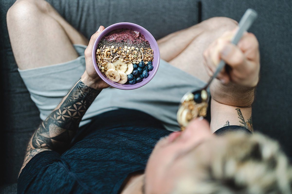 A man is laying on a couch eating a bowl of cereal.