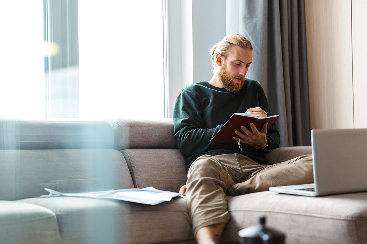 A man is sitting on a couch with a laptop and a notebook.