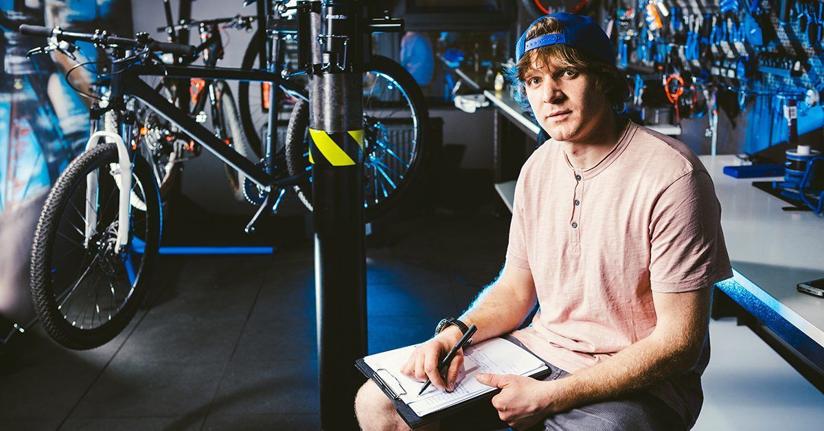 A man is sitting in front of a bicycle in a garage holding a clipboard.