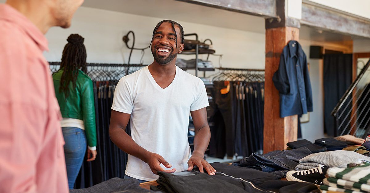 A man is standing behind a counter in a clothing store talking to a customer.