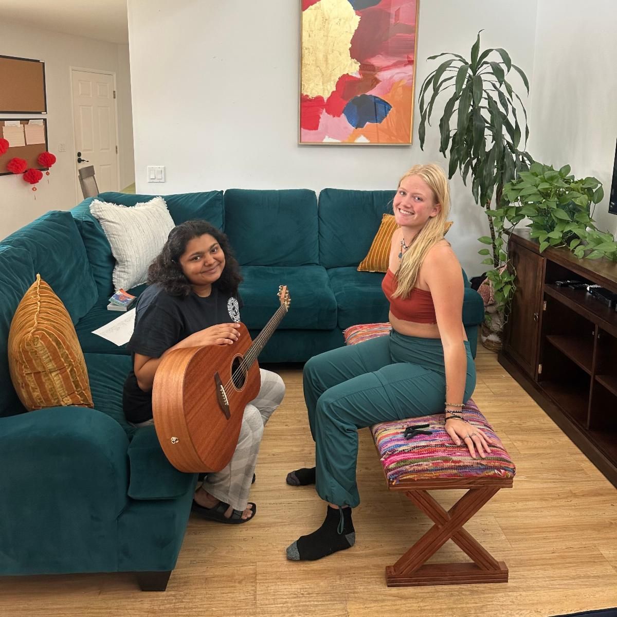 Two women in a living room, one holding a guitar, the other seated on a footstool, smiling.