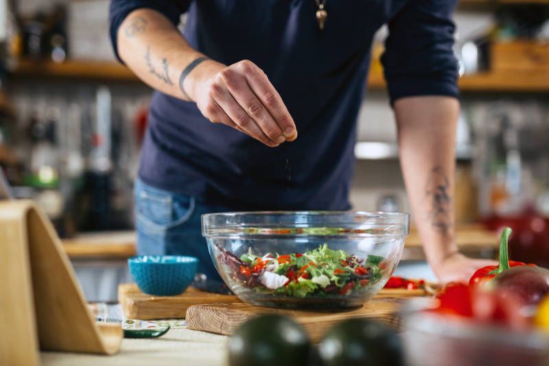 A man is adding salt to a salad in a bowl.