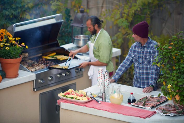 Two men are cooking food on a grill in an outdoor kitchen.