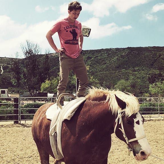 A boy wearing a california shirt stands on top of a brown horse