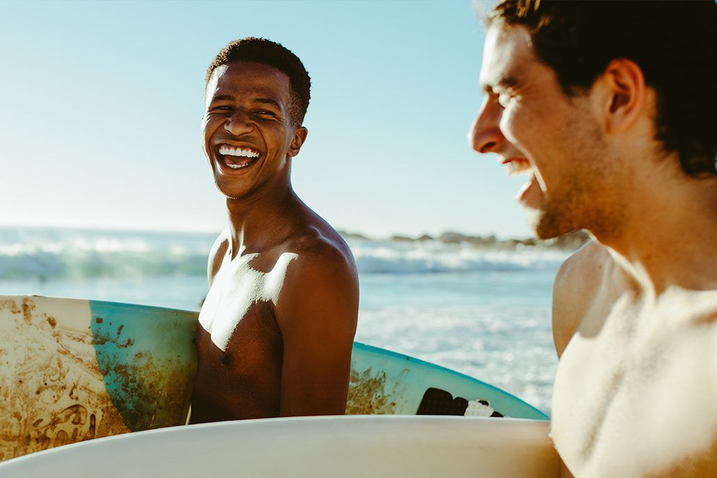 Two men are holding surfboards on the beach and smiling.