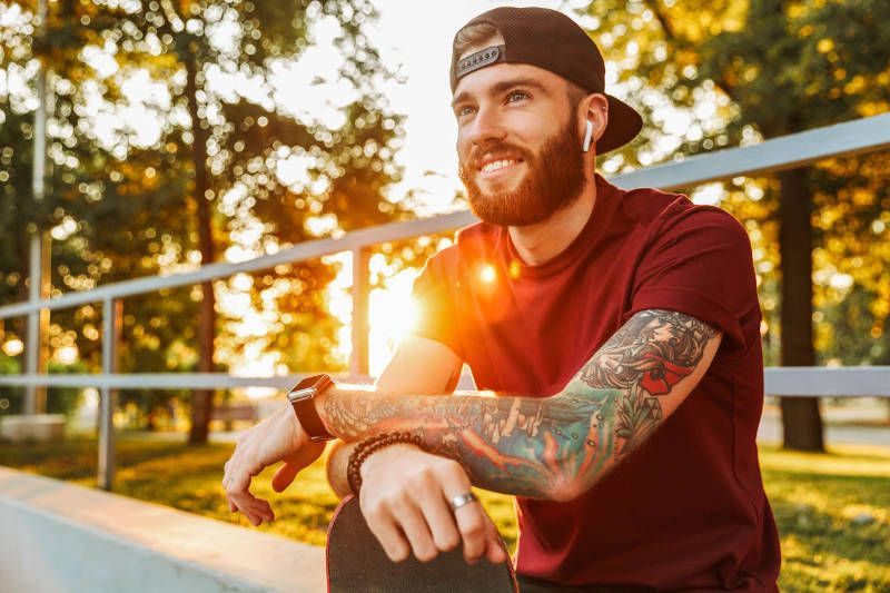 A man with a beard and tattoos is sitting on a skateboard in a park.