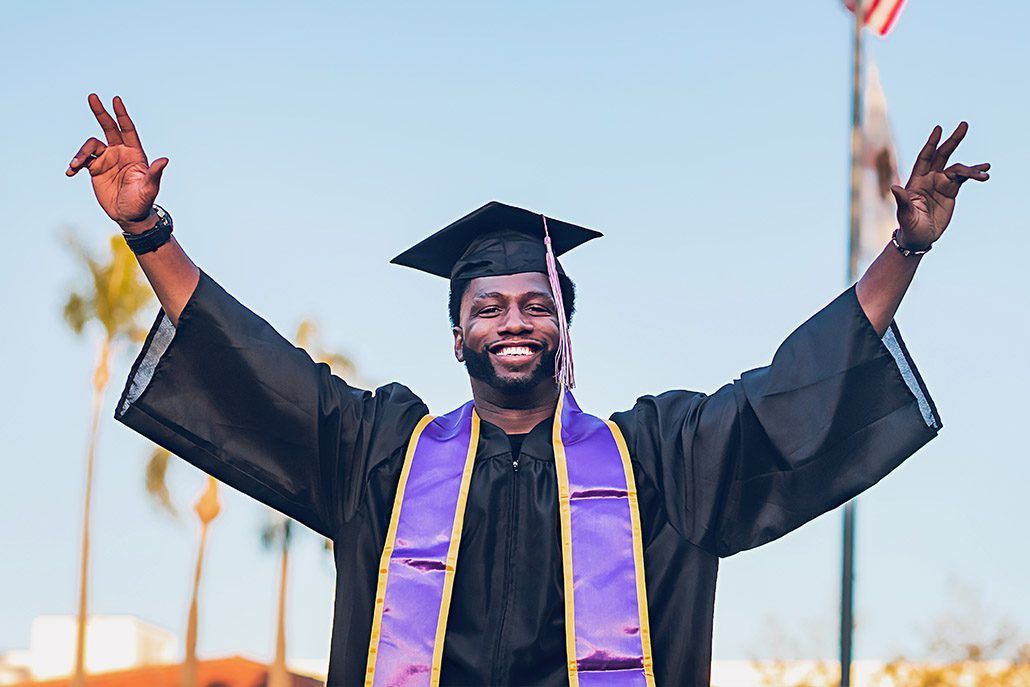 A man in a graduation cap and gown is standing with his arms outstretched.