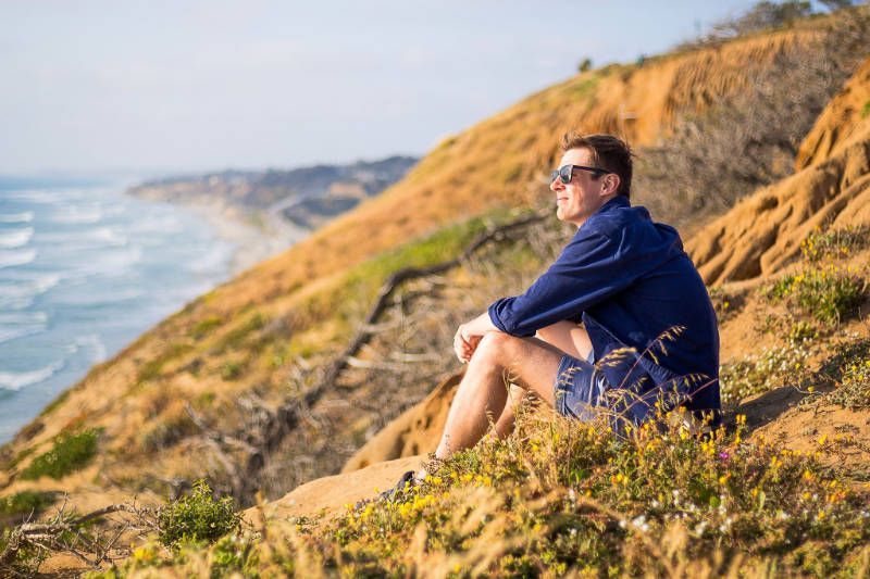 A man is sitting on top of a cliff overlooking the ocean.